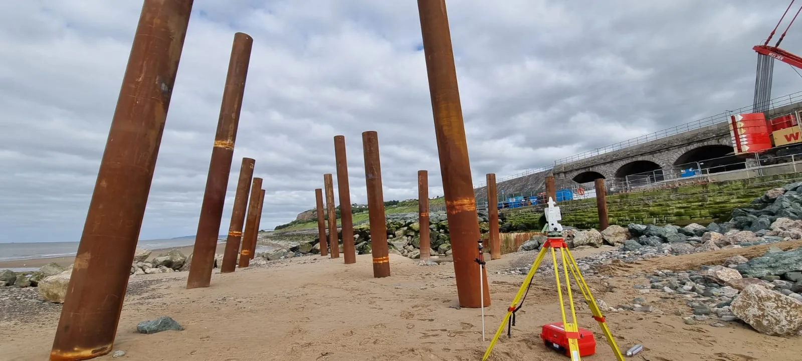 Steel piles on a beach beside a sea wall, with a total station on a yellow tripod