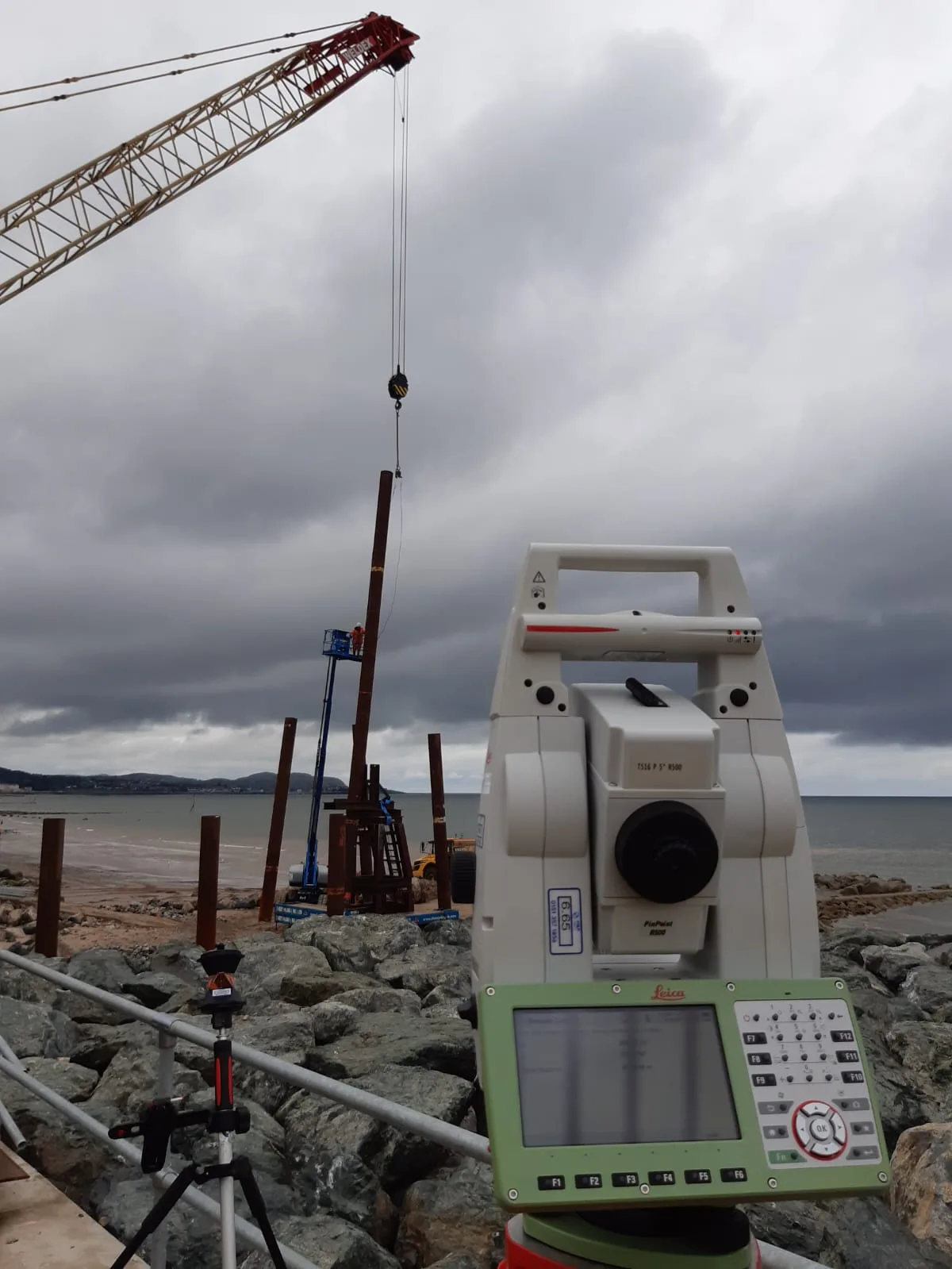 Robotic total station on rock armour, monitoring a steel pile being lifted into place by a crane on the coast