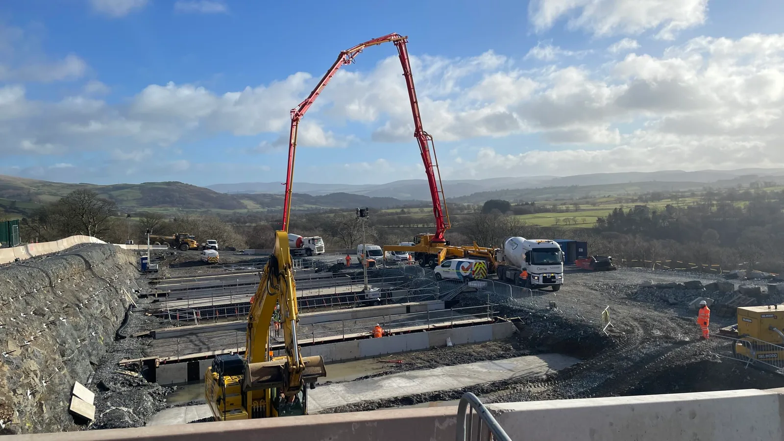 Concrete pump boom reaching over formwork in a large excavation, with mixer lorries and hills beyond