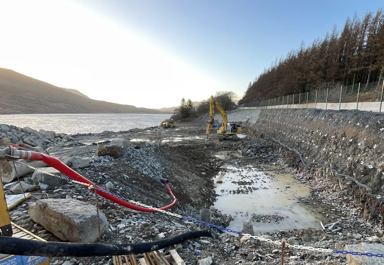 Excavators working in a deep cutting beside a reservoir, with soil nail plates visible across the rock face