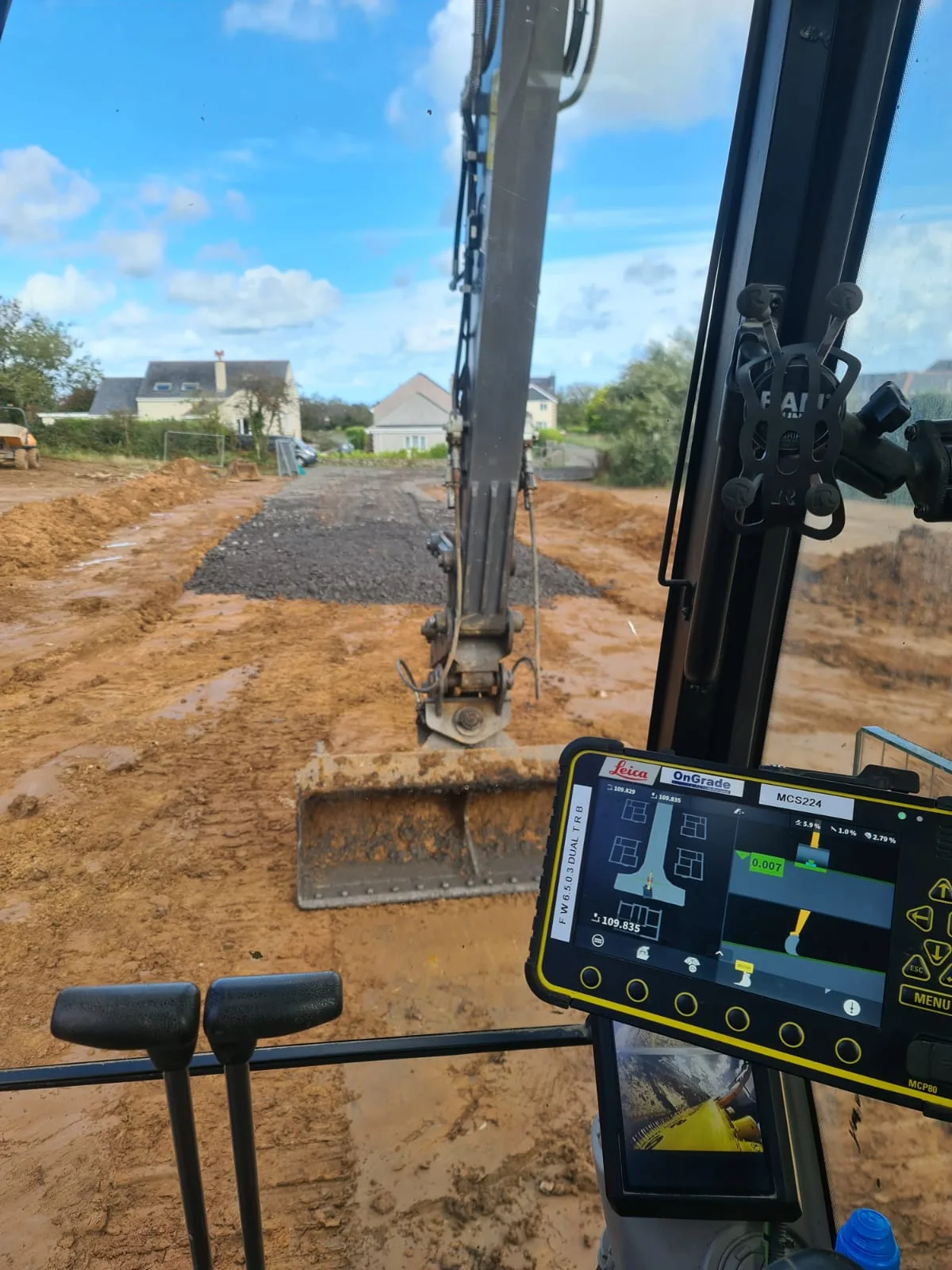 View from an excavator cab over a levelled plot, with the machine control screen in view