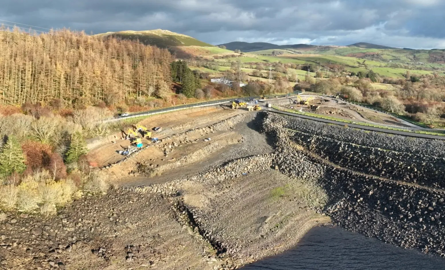Aerial view of earthworks on a reservoir embankment, with excavators, dump trucks and rock armour, and autumn forest and hills beyond